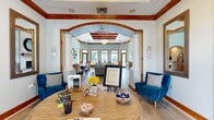This single-floor amenity space offers a thoughtfully designed layout spanning a generous footprint, combining social, recreational, and fitness functions under one roof. The main living area immediately draws attention with its soaring coffered ceiling accented by rich wood beams, a statement chandelier, and a wall of multi-pane windows that flood the room with natural light. A stone fireplace with a decorative mirror above serves as a focal point, complemented by light hardwood flooring and soft sage-toned walls throughout.

Elegant arched doorways framed in warm wood trim connect the living room to adjacent spaces, including a welcoming entry area with high ceilings and a separate dining and kitchen zone. The dining and lounge area features warm wood panel wainscoting, tile flooring, white upholstered seating, and a shuffleboard table, providing a versatile gathering space. A connected kitchen area with recessed lighting and outdoor-facing doors adds convenience.

The dedicated exercise room is well-equipped with cardio machines, strength equipment, a dumbbell rack, wall-to-wall mirrors, and natural light from a side window, creating a functional and motivating fitness environment.