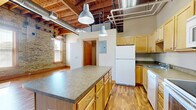This studio unit at 315 Main Ave in Moorhead, MN offers a thoughtfully designed open-concept layout that blends industrial loft character with functional living. The space is defined by striking exposed wooden beam ceilings with visible ductwork and piping, lending an authentic urban warehouse aesthetic. Original brick walls with wood-trimmed windows flood the living area with natural light, creating a warm and inviting ambiance throughout.

The kitchen is a central highlight, featuring a large island with a laminate countertop, light wood cabinetry, and a full suite of white appliances including a refrigerator, electric range, over-the-range microwave, and dishwasher. Industrial-style pendant lights hang above the island, reinforcing the loft character. The kitchen flows seamlessly into the open living area, where rich wood-toned laminate flooring extends throughout the entire unit.

The living space benefits from multiple windows along the exposed brick wall, allowing abundant natural light to pour in. The bathroom includes a clean, white walk-in shower stall. The unit also features a dedicated closet area with a hanging rod for storage. The overall design balances the rugged, raw charm of the building's industrial heritage with comfortable, modern finishes suitable for everyday living.