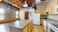 This studio unit at 315 Main Ave in Moorhead, MN offers a thoughtfully designed open-concept layout that blends industrial loft character with functional living. The space is defined by striking exposed wooden beam ceilings with visible ductwork and piping, lending an authentic urban warehouse aesthetic. Original brick walls with wood-trimmed windows flood the living area with natural light, creating a warm and inviting ambiance throughout.

The kitchen is a central highlight, featuring a large island with a laminate countertop, light wood cabinetry, and a full suite of white appliances including a refrigerator, electric range, over-the-range microwave, and dishwasher. Industrial-style pendant lights hang above the island, reinforcing the loft character. The kitchen flows seamlessly into the open living area, where rich wood-toned laminate flooring extends throughout the entire unit.

The living space benefits from multiple windows along the exposed brick wall, allowing abundant natural light to pour in. The bathroom includes a clean, white walk-in shower stall. The unit also features a dedicated closet area with a hanging rod for storage. The overall design balances the rugged, raw charm of the building's industrial heritage with comfortable, modern finishes suitable for everyday living.