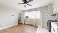 This single-floor studio is designed for efficient, comfortable living with a clean and functional layout. The main living area features light wood laminate flooring that extends throughout, neutral gray-toned walls, and a ceiling fan with a light fixture that provides both airflow and ambient lighting. Natural light filters in through a set of windows fitted with horizontal blinds, creating a bright and airy atmosphere. A wall-mounted heating and cooling unit, along with a window air conditioning unit, ensures year-round climate control.

The open kitchen area transitions seamlessly from the living space, distinguished by white tile flooring, white shaker-style cabinetry with a subway tile backsplash, a single-basin sink, and a full-size refrigerator. Adjacent to the kitchen, a set of built-in open shelving units offers generous storage and display space, adding a practical yet visually appealing architectural detail to the unit.

The bathroom is neatly appointed with white hexagonal floor tile, a white vanity with a single-basin sink and matte black fixtures, a rectangular mirror with a navy frame, and a full-size soaking tub with a shower, tiled in classic white square tiles. A grab bar and stylish pendant lighting complete the thoughtfully detailed bathroom space.