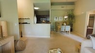 This single-story residence features an open-concept layout that seamlessly connects the living, dining, and kitchen areas into one cohesive living environment. The living room is finished with plush neutral carpeting and painted in warm, earthy tones that create a calm and welcoming atmosphere throughout.

The kitchen is partially separated from the main living space by a half-wall pass-through, offering both visual connection and functional division. It is equipped with dark espresso cabinetry, stainless steel appliances including a microwave, and light-colored countertops that contrast attractively against the darker cabinetry. A decorative white vase accents the pass-through ledge, adding a touch of style.

Adjacent to the kitchen, the dining area is defined by a pendant light fixture that hangs overhead, illuminating a wooden dining table surrounded by upholstered chairs. Framed artwork adorns the olive-green accent wall behind the dining space, complementing the lush indoor plant nearby.

The overall color palette—combining warm beiges, sage greens, and deep browns—lends the space a grounded, nature-inspired ambiance suited for everyday living.