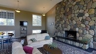 This single-story home centers around a striking living room defined by its soaring vaulted ceilings and a commanding floor-to-ceiling stone fireplace. Constructed from natural fieldstone in varied earthy tones, the fireplace serves as the room's focal point, spanning the full height of the wall and adding a rustic yet refined character to the space. The vaulted ceiling amplifies the sense of openness and volume throughout the living area. Natural light enters through multiple windows, including a prominent upper window that draws light deep into the room and offers views of surrounding trees, enhancing the connection to the outdoors. The living room is furnished with a plush, cream-colored sofa set accented by colorful throw pillows, arranged around a wooden oval coffee table adorned with a potted plant. A patterned area rug anchors the seating arrangement, adding warmth and texture underfoot. A media console with a flat-screen television occupies one corner, complemented by shelving for storage. The warm, taupe-toned walls and thoughtful furnishings create a cozy, welcoming ambiance suited for relaxed everyday living.