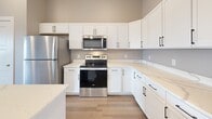 Located at The Elements in Rapid City, South Dakota, this well-appointed single-floor unit blends contemporary design with functional living. The open-concept kitchen is a standout feature, outfitted with crisp white shaker cabinetry, quartz countertops, stainless steel appliances including a range, microwave, and refrigerator, and a central island with a built-in sink and pendant lighting overhead. Wood-look luxury vinyl flooring runs throughout the main living areas, complementing the neutral color palette and clean lines throughout.

The living area is generously proportioned, featuring exposed metallic ductwork that adds an industrial character to the space, along with a ceiling fan and ample room for furnishings. A sliding glass door opens to a private balcony with railing, offering open views of the surrounding Rapid City landscape.

The bedrooms are carpeted for comfort, with one featuring dual bi-fold closet doors providing abundant storage, and another with a spacious walk-in closet. Two full bathrooms serve the unit — one with a double-sink vanity featuring white cabinetry and quartz countertops, and another with a single vanity, full mirror, and shower/tub combination. An in-unit laundry area is also present, adding everyday convenience.
