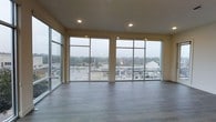 This single-floor space makes a striking first impression with its expansive, open layout and thoughtfully modern design. The most defining feature is the dramatic wall of floor-to-ceiling windows that spans the main living area, flooding the interior with abundant natural light and providing panoramic views of the surrounding cityscape and beyond. The large-format window panels are framed in sleek metal, adding a clean, contemporary architectural element to the space. The smooth, light-toned walls complement the warm, wood-look luxury vinyl plank flooring, which runs throughout and adds both texture and durability to the interior. Recessed lighting is integrated into the ceiling, offering a polished, unobtrusive source of illumination that enhances the space during evening hours. The elevated position of the space amplifies the sense of openness, allowing natural light to pour in from multiple angles. The overall ambiance is airy, minimalist, and versatile, well-suited for a variety of uses. The combination of high ceilings, generous glazing, and clean finishes creates an environment that feels both functional and refined, with the ever-changing outdoor scenery serving as a natural backdrop.