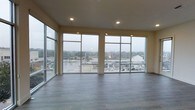 This single-floor space makes a striking first impression with its expansive, open layout and thoughtfully modern design. The most defining feature is the dramatic wall of floor-to-ceiling windows that spans the main living area, flooding the interior with abundant natural light and providing panoramic views of the surrounding cityscape and beyond. The large-format window panels are framed in sleek metal, adding a clean, contemporary architectural element to the space. The smooth, light-toned walls complement the warm, wood-look luxury vinyl plank flooring, which runs throughout and adds both texture and durability to the interior. Recessed lighting is integrated into the ceiling, offering a polished, unobtrusive source of illumination that enhances the space during evening hours. The elevated position of the space amplifies the sense of openness, allowing natural light to pour in from multiple angles. The overall ambiance is airy, minimalist, and versatile, well-suited for a variety of uses. The combination of high ceilings, generous glazing, and clean finishes creates an environment that feels both functional and refined, with the ever-changing outdoor scenery serving as a natural backdrop.