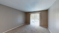 This single-floor residence offers a practical and comfortable layout designed for everyday living. The living area is generously sized, featuring neutral-toned carpet and warm taupe walls that create a welcoming atmosphere. A sliding glass door fitted with vertical blinds fills the space with natural light and provides direct outdoor views, while a front entry door with hardwood-style flooring transitions seamlessly into the carpeted living space. The open-concept design connects the living area to the kitchen via a pass-through counter with white cabinetry, offering convenient sight lines throughout. The kitchen is equipped with black appliances, including a refrigerator, electric coil range, and an overhead range hood, complemented by tile backsplash and light wood-style flooring. A hallway with a ceiling fan leads to the bedroom, which is carpeted and features a window with horizontal blinds that allows natural light to filter in. The bathroom is well-finished, showcasing a white vanity with granite-style countertop, undermount sink, and brushed nickel fixtures. A large mirror with overhead vanity lighting, wood-style flooring, and a tub-shower combination round out this clean and functional bathroom. Throughout the unit, consistent neutral tones and clean lines contribute to a cohesive, move-in-ready aesthetic.