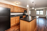 Located at 5485 Mills Civic Pkwy in West Des Moines, Iowa, this single-floor unit offers a well-organized layout that maximizes its compact footprint. The open-concept kitchen and living area form the heart of the space, where rich hardwood-style flooring runs throughout, creating a warm and cohesive aesthetic. The kitchen is thoughtfully appointed with warm-toned wood cabinetry extending to the upper walls for generous storage, dark countertops, a full suite of black appliances — including a refrigerator, electric range, built-in microwave, and dishwasher — and a central island with pendant lighting overhead. The island provides additional prep space and serves as a natural divider between the kitchen and the open living area. The living space is bright and airy, with multiple windows offering views of the surrounding West Des Moines cityscape and allowing natural light to fill the room. The bathroom features tile flooring, a wood-cabinet vanity with dark countertop, and an in-unit stacked washer and dryer conveniently accessible from the same space. A spacious walk-in closet with full wire shelving and hanging rods provides ample storage. The overall ambiance is clean, modern, and functional.