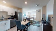 Located at 1525 W Arrowood Rd in Charlotte, NC, this single-floor suite offers a thoughtfully designed layout that balances comfort and functionality. The open-concept living and dining area is anchored by light wood-look flooring, neutral walls, and ample natural light streaming through window blinds. A compact dining table with teal upholstered chairs sits adjacent to the kitchen, creating a seamless transition between spaces. The fully equipped kitchen features white cabinetry, granite-style countertops, stainless steel appliances including a refrigerator, electric range, microwave, and dishwasher — providing everything needed for everyday cooking. The living area offers a comfortable sofa, accent chairs, and decorative blue-toned artwork that carries a cohesive coastal aesthetic throughout the space. The bedroom is a serene retreat with a king-sized bed, matching nightstands, wall-mounted TV, a walk-in closet, and a window with outdoor views. The bathroom includes a granite vanity, a glass-enclosed walk-in shower, and a convenient in-unit washer tucked into a dedicated nook. Clean finishes and a consistent color palette give the entire space a modern, welcoming feel.
