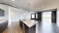 This thoughtfully designed single-floor space showcases a seamless blend of modern and industrial aesthetics. The open-concept layout unifies the kitchen and living areas, creating a spacious and airy atmosphere. Exposed silver ductwork runs across the ceiling, adding an industrial character that complements the contemporary finishes throughout.

The kitchen is anchored by a generously sized island featuring a light quartz countertop, an undermount double sink with a modern arc faucet, dark espresso cabinetry, and a stainless steel dishwasher. Pendant lights with metallic accents hang above the island, providing focused task lighting and a stylish focal point.

Wide-plank hardwood flooring in a warm brown tone flows continuously throughout the space, enhancing its cohesive and polished look. The living area is bathed in natural light from large windows and a glass door that opens to an outdoor balcony, offering views of the surrounding landscape. Dark accent walls near the balcony entrance create a striking contrast against the bright white walls and ceilings. Recessed lighting supplements the natural light, ensuring the space remains well-lit throughout the day. The overall ambiance is clean, contemporary, and inviting.