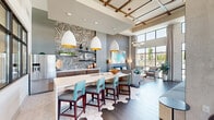 Located at 2511 Watermark Ter in Elgin, IL, this thoughtfully designed community clubhouse delivers a modern, resort-inspired atmosphere across a generously sized single-floor layout. The space is anchored by a soaring double-height ceiling with exposed wood beam detailing and expansive floor-to-ceiling windows that flood the interior with natural light and offer views of the outdoor pool area and surrounding grounds.

The living area showcases a striking floor-to-ceiling wood-clad fireplace column with a sleek linear gas fireplace and wall-mounted television, serving as the room's dramatic focal point. Dark-toned wood plank flooring runs throughout, complementing the warm neutral and grey tones of the walls. Curated lounge seating—including a vibrant blue velvet sofa, light grey armchairs, and a natural wood round coffee table—creates an inviting and contemporary gathering space. A bold geometric textured wall accent and a triptych of abstract aqua artwork add artistic character.

A dedicated recreation area features a full-sized billiards table beneath pendant lighting, alongside built-in banquette seating and café-style dining. Statement pendant and chandelier lighting fixtures enhance the sophisticated ambiance throughout. Glass double doors provide seamless access to the outdoor pool terrace, further extending the social and recreational experience of this well-appointed clubhouse.