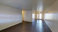 This single-story space showcases a open main living area defined by rich, dark hardwood floors that run throughout, creating a warm and cohesive aesthetic. The textured popcorn ceiling adds a classic touch overhead, while a ceiling fan is centrally positioned to promote comfortable air circulation. Natural light filters generously into the space through a set of double front doors fitted with glass panels, brightening the interior and lending an inviting atmosphere. The walls are painted in a clean, neutral white, providing a versatile backdrop suitable for a variety of décor styles. A louvered door and additional closet or utility access doors are visible along the back wall, offering practical built-in storage solutions. A floor vent indicates a central HVAC system for climate control. The layout appears open and functional, with clear sightlines across the main living area toward the entryway. The overall ambiance is clean, simple, and move-in ready, with foundational finishes that balance everyday practicality with understated style.
