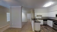 This single-floor residence offers a well-planned layout that balances open communal spaces with private retreats. The kitchen is a standout feature, showcasing white shaker-style cabinetry, dark countertops, a classic subway tile backsplash, and stainless steel appliances. A peninsula with additional counter space and storage extends into the living area, creating a natural divide between the kitchen and the carpeted living room. Crown molding throughout adds a refined architectural touch, lending the space an elevated, polished character. The flooring transitions from rich, dark hardwood-style planks in the kitchen and hallway to plush carpet in the living and bedroom areas, offering both practicality and comfort underfoot. A hallway connects the main living area to the private bedroom, which features a ceiling fan, carpeted flooring, and a window that allows natural light to filter in. A door along the hallway provides access to an additional room or storage space, enhancing the overall functionality of the layout. The neutral color palette of warm beige and crisp white tones throughout creates a clean, inviting ambiance that is easy to personalize. Overall, the space presents a practical and comfortable single-level living environment with thoughtful design details.