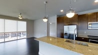This contemporary single-floor Dallas residence showcases a thoughtfully designed open-concept layout that seamlessly connects the kitchen and living areas. The gourmet kitchen serves as the centerpiece of the space, featuring a large island topped with striking gold-veined granite countertops and an undermount sink with a professional-style faucet. Pendant lighting with polished chrome fixtures hangs above the island, casting a warm glow over the workspace. Rich, dark espresso cabinetry lines the kitchen walls, providing generous storage and a bold contrast against the lighter elements of the space. A stainless steel French door refrigerator with a bottom freezer adds a modern, high-end touch to the culinary area. Dark hardwood floors flow throughout, grounding the space with warmth and elegance. The open living area receives natural light through a large window, creating a bright and welcoming atmosphere. Clean white walls and recessed lighting complement the overall modern aesthetic, while an open doorway hints at additional rooms beyond. The combination of high-quality materials, thoughtful lighting, and cohesive design elements makes this Dallas residence a refined and functional living environment.