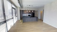 This single-floor space showcases a thoughtfully designed open-concept layout that seamlessly connects the kitchen and living areas. The kitchen is a standout feature, boasting dark wood cabinetry, a subway tile backsplash, and a large central island that provides generous counter space and serves as a natural focal point for the room. Black appliances, including a refrigerator, stove, and microwave, lend a cohesive, contemporary aesthetic. Pendant lights hang above the island, adding warm, focused illumination, while a track lighting system overhead provides additional flexibility. An exposed industrial-style ventilation duct runs along the ceiling, contributing an urban loft character to the space. Wide-plank light hardwood flooring extends throughout the open living area, creating a sense of warmth and continuity. Large windows fitted with horizontal blinds allow abundant natural light to flood the interior, enhancing the airy, open atmosphere. A hallway visible in the background suggests access to additional private rooms, rounding out the functional layout of this modern, well-appointed space.