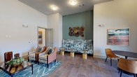 This thoughtfully designed single-floor community lounge offers a welcoming and visually dynamic environment that blends comfort with contemporary style. The space features soaring ceilings that enhance the open, airy feel throughout, while large tile flooring in warm earth tones grounds the room with a sense of understated elegance.

The main seating area is anchored by a boldly patterned sofa set against a deep teal accent wall, adorned with an eye-catching circular wood-slice wall sculpture. Complementing the sofa are two mixed-pattern armchairs arranged over a vibrant blue geometric area rug, with polished gold side tables adding a refined metallic accent throughout.

A set of large botanical canvas prints decorates an adjacent wall, further reinforcing the nature-inspired aesthetic. This seating zone flows naturally into a more intimate dining nook, furnished with a round blue-toned table and cognac leather chairs. Floor-to-ceiling glass doors in this area open to an outdoor balcony, inviting natural light and providing views of the surrounding exterior.

Curated artwork is displayed throughout the space, including a warm landscape painting, contributing to a gallery-like atmosphere. A connecting corridor leads further into the building, suggesting additional amenities beyond. Overall, the space presents a polished, cohesive design that balances social functionality with distinctive interior character.
