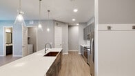 This well-appointed two-story home offers 2 bedrooms and 3 bathrooms across two floors, with the expansive main level accounting for the majority of the living space and an upper level providing additional area. The open-concept kitchen and living room serve as the heart of the home, showcasing gray shaker-style cabinetry, white quartz countertops, a large island with a double undermount sink, stainless steel appliances including a refrigerator with ice dispenser, electric range, and microwave, plus a built-in wine rack. Elegant glass pendant lights illuminate the island, while light wood-look flooring and recessed lighting create a bright, modern atmosphere throughout. The living area is spacious and airy, featuring a ceiling fan and large windows that invite natural light. The primary bedroom connects directly to a well-designed ensuite bathroom with a dual-sink vanity, gray cabinetry, white quartz countertops, and framed mirrors, all finished in a cohesive neutral palette. Soft carpet flooring adds comfort to the bedrooms. A dedicated laundry room houses a full-size washer and dryer with overhead shelving. The attached two-car garage provides ample storage and parking. Outdoors, a landscaped green space with mature trees and an iron fence offers a pleasant setting.