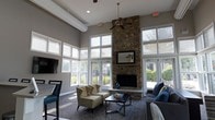 This single-story lounge space makes a striking impression with its soaring ceiling height, creating an open and airy atmosphere throughout. The room's most commanding feature is a floor-to-ceiling stacked stone fireplace, which serves as a natural focal point and anchors the space with a warm, rustic character. Flanking the fireplace, a mounted flat-screen television adds a functional element to the gathering area.

Large windows wrap around the perimeter, filling the room with generous natural light and offering clear views of the surrounding outdoor landscape. Transom windows positioned near the ceiling further enhance the sense of height and brightness. A ceiling fan helps regulate airflow throughout this expansive volume.

The seating arrangement includes a pair of cushioned armchairs alongside a low-profile coffee table, complemented by a plush sofa adorned with navy accent pillows, creating a comfortable and inviting layout. The combination of textured stone, neutral wall tones, and contemporary furnishings strikes a balance between casual comfort and polished design, making this a welcoming communal retreat.