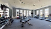 This fitness center is a thoughtfully designed, single-floor facility built to support a wide range of workout routines. The space features neutral gray walls, durable carpet flooring, and a clean, modern aesthetic throughout. Recessed lighting and multiple ceiling fans ensure the environment remains well-lit and comfortable during any workout session.

Large windows line the walls, flooding the interior with abundant natural light and offering views of the surrounding community. A wall-mounted flat-screen television provides entertainment during cardio sessions. Full-length mirrors on one wall visually expand the space and assist with form and technique.

The cardio section is well-appointed with multiple treadmills, an elliptical, a stair climber, and a stationary spin bike. The strength training area includes a rack of dumbbells, adjustable workout benches, a multi-functional cable machine, and a dedicated resistance machine. A medicine ball rack stocked with a variety of weighted balls and a stability ball rounds out the functional training options.

An interior window and glass door connect the fitness center to an adjacent common area, promoting a sense of openness and easy accessibility. The overall layout is spacious and uncluttered, allowing users to move freely between equipment zones and enjoy a well-organized, professional-grade workout environment.