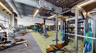 Located at 419 W Sixth St in Covington, Kentucky, this fitness facility combines an industrial aesthetic with a comprehensive array of workout equipment. The single-floor space features soaring ceilings with exposed ductwork, structural columns, and linear pendant lighting that illuminate the open floor plan. Patterned carpet tiles in green and gray tones define distinct workout zones throughout the space.

The strength training area is prominently anchored by a TRX Training Zone, complete with multi-functional rig systems, barbells, and weight plate storage. Multiple power racks and benches are arranged in an orderly row, providing ample space for free weight training. An extensive dumbbell rack lines one wall, backed by floor-to-ceiling mirrors that visually expand the space.

A dedicated functional training zone occupies a central area, outfitted with medicine ball racks, battle ropes, kettlebells, and resistance bands stored on vertical racks. Large front-facing windows along one wall allow natural light to enter and offer street-level views. Cardio equipment, including treadmills and upright machines, is positioned near the windows.

A utility area toward the rear houses exercise mats, stability balls, and additional accessories, alongside exposed brick accents that add warmth to the otherwise modern, industrial interior.