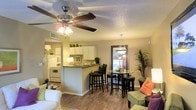 This single-floor residence showcases an open-concept design that seamlessly integrates the living, dining, and kitchen areas into one cohesive, functional space. The living area is anchored by a ceiling fan with a built-in light fixture, providing both comfort and illumination, while warm wood-style flooring flows throughout, lending a welcoming and unified aesthetic.

The kitchen features white cabinetry, a white appliance suite including a refrigerator and range, and a peninsula with a dark countertop that offers bar-style seating with tall black stools. This peninsula serves as a natural divider between the kitchen and the rest of the open living space, making it ideal for casual dining or entertaining.

Adjacent to the kitchen, the dining area accommodates a round glass-top table with dark chairs, accented by a pendant light fixture overhead. A large mirror on the dining room wall enhances the sense of space and reflects natural light.

The living area features comfortable seating with colorful accent pillows, adding pops of color against the neutral wall tones. A tall indoor plant near the dining space introduces a natural element, contributing to the overall warm and inviting ambiance of the home.