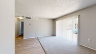 This single-floor residence offers a thoughtfully arranged layout comprising a living room, kitchen, bedroom, bathroom, and closet. The living room is a bright, open area featuring a combination of light wood-style flooring and plush carpet, neutral gray walls, a textured ceiling, and a large sliding glass door dressed with vertical blinds that invites generous natural light and outdoor views. The entry area transitions seamlessly between the carpeted living space and the hard-surface flooring leading into the kitchen. The kitchen is efficiently designed with white cabinetry, white appliances including a range, refrigerator, and dishwasher, a single-basin sink, and light wood-style flooring throughout. The spacious primary bedroom is carpeted with neutral tones, features a window with natural light, and connects to a walk-in closet equipped with built-in wire shelving for organized storage. The full bathroom includes a tiled tub-shower combination, a toilet, vanity with countertop, and wood-style flooring. Clean lines, a consistent neutral color palette, and practical finishes contribute to a functional and comfortable living environment throughout.