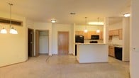 This single-floor residence offers a functional and welcoming open-concept layout that seamlessly connects the living area to the kitchen. The carpeted living space flows into a tile-floored kitchen, creating a clear distinction between the two zones while maintaining an open and airy feel. The kitchen is equipped with light maple cabinetry, black appliances including a refrigerator, microwave, and range, and is anchored by a white breakfast bar peninsula that doubles as a casual dining or entertaining surface. Two pendant lights hang above the bar, adding a warm, focused glow to the kitchen area, which is further illuminated by an overhead light fixture. A central entry door with warm wood tones provides a welcoming focal point at the back of the space. To the left, an open closet offers practical storage with visible shelving and a hanging rod, ideal for coats or additional organization. The neutral color palette throughout — from the beige carpet to the light walls and cabinetry — creates a clean, versatile backdrop that is easy to personalize. The layout is efficient and well-organized, making the most of the available single-level floor plan.