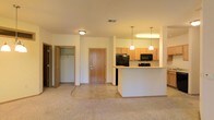 This single-floor residence offers a functional and welcoming open-concept layout that seamlessly connects the living area to the kitchen. The carpeted living space flows into a tile-floored kitchen, creating a clear distinction between the two zones while maintaining an open and airy feel. The kitchen is equipped with light maple cabinetry, black appliances including a refrigerator, microwave, and range, and is anchored by a white breakfast bar peninsula that doubles as a casual dining or entertaining surface. Two pendant lights hang above the bar, adding a warm, focused glow to the kitchen area, which is further illuminated by an overhead light fixture. A central entry door with warm wood tones provides a welcoming focal point at the back of the space. To the left, an open closet offers practical storage with visible shelving and a hanging rod, ideal for coats or additional organization. The neutral color palette throughout — from the beige carpet to the light walls and cabinetry — creates a clean, versatile backdrop that is easy to personalize. The layout is efficient and well-organized, making the most of the available single-level floor plan.