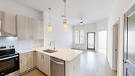 This well-appointed one-bedroom, one-bathroom residence at 2800 Milan Ct in Birmingham, Alabama offers a thoughtfully designed layout across a single floor. The open-concept kitchen serves as a focal point, featuring light wood cabinetry, white subway tile backsplash, stainless steel appliances including a side-by-side refrigerator with ice maker and an electric range, and a peninsula with a built-in sink and light quartz countertops. Pendant lighting above the peninsula adds a contemporary touch, while warm hardwood-style flooring flows throughout the main living areas. Distinctive slate-blue double doors and interior doors provide a stylish contrast to the crisp white walls throughout the home. The bedroom is a calm, comfortable retreat finished with plush neutral carpet and a window that invites natural light. The bathroom is sleek and modern, showcasing a floating wood-toned vanity with white countertop, a large mirror with bar lighting, and the same warm hardwood-style flooring carried in from the main areas. The overall aesthetic is clean and contemporary, with a neutral palette and quality finishes that create a cohesive and inviting atmosphere throughout the space.