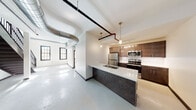 This two-story space showcases a compelling blend of industrial design and contemporary upgrades. The main level centers around an open-concept living and kitchen area, where polished concrete floors reflect natural light streaming through large, black-framed windows. Exposed silver ductwork runs prominently across the ceiling, reinforcing the industrial aesthetic, while pendant lighting adds a modern touch above the kitchen island. The island features a sleek white quartz countertop with an undermount sink, contrasting against a dark metal base. The kitchen is equipped with stainless steel appliances, rich dark-toned cabinetry reaching to the ceiling, and track lighting overhead. A bold black metal staircase leads to the upper level, where carpeted flooring and an open railing create a loft-like landing. The upper floor houses additional rooms, including carpeted bedrooms with black-framed windows that invite natural light, and bathrooms on both levels finished with light wood-look flooring, dark vanities, white quartz countertops, and large mirrors. Exposed piping accents carry the industrial theme throughout. The overall ambiance is modern and urban, with clean white walls balancing the raw architectural elements.