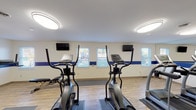 Located at 6615 Fountains Blvd in West Chester, Ohio, this single-floor fitness center offers a well-equipped and thoughtfully designed workout space. The interior is finished with light hardwood-style flooring that runs throughout the room, creating a clean and polished aesthetic. A bold blue horizontal stripe accents the white walls, giving the space a cohesive, energetic visual identity.

The facility is outfitted with a comprehensive selection of exercise equipment, including multiple treadmills, elliptical cross-trainers, a recumbent bike, a multi-function strength training tower, and free weights. Equipment mats are placed beneath each station, providing both safety and organization. A large wall mirror along one side of the room enhances the sense of space and allows users to monitor their form during workouts.

Multiple flat-screen televisions are mounted on the walls throughout the room, keeping members entertained during cardio sessions. Natural light filters in through several windows fitted with blinds, offering views of the surrounding outdoor area. Overhead flush-mount and oval lighting fixtures provide bright, even illumination across the entire space. The room also includes a designated open area along one wall, suitable for stretching, mat work, or floor exercises, making this fitness center a versatile and functional space for a full range of workout routines.