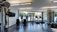 This single-floor fitness center is thoughtfully designed to provide an open, motivating environment for exercise and wellness. The space is defined by its expansive floor-to-ceiling windows that line the far wall, allowing an abundance of natural light to pour in while offering clear views of the surrounding outdoor greenery and streetscape — creating a refreshing, energizing atmosphere.

The interior features a clean, modern aesthetic with white walls, a smooth light-toned ceiling equipped with industrial-style linear pendant lighting, and a durable dark speckled flooring that is both practical and visually grounding. A wall-mounted flat-screen television is positioned prominently on one side, allowing members to stay entertained or informed during their workouts.

The fitness floor is outfitted with a variety of cardio equipment, including multiple treadmills, elliptical cross-trainers, and a stationary cycle, all arranged to maximize usable space while maintaining comfortable circulation between machines. Double glass entry doors provide direct access from the exterior, contributing to the facility's bright and welcoming character. Overall, the space strikes a balance between functional design and an inviting, light-filled ambiance ideal for daily fitness routines.