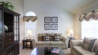 This single-story space showcases a thoughtfully designed living area that combines comfort and classic style. The room's most striking architectural feature is its vaulted ceiling, which rises dramatically to create an expansive, airy feel. An arched window with a decorative fabric valance in a floral toile pattern allows natural light to filter softly into the space, complementing the neutral, light-toned walls throughout.

The living area is furnished with a generously sized, beige upholstered sofa adorned with an assortment of plaid and patterned accent pillows in earth tones, adding visual texture and warmth. A matching armchair is partially visible, suggesting a well-composed seating arrangement. Flanking the sofa are dark wood side tables, each topped with classic table lamps that provide warm ambient lighting.

A sleek, dark-framed coffee table serves as the focal point of the seating arrangement, displaying a decorative bowl and reading material. Above the sofa, four uniformly framed black-and-white photographic prints are arranged in a symmetrical grid, contributing a refined, cohesive aesthetic. Neutral carpet flooring ties the room together, reinforcing the overall warm and inviting ambiance of the space.