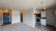 This single-floor unit offers a practical and efficient layout designed to maximize functionality. The living area is carpeted and open, featuring a textured ceiling, neutral gray walls, and a large window that allows natural light to fill the space while providing views of the surrounding neighborhood. A baseboard heater runs along the exterior wall, and a wall-mounted AC unit ensures year-round comfort.

The kitchen is semi-open and connects seamlessly to the living space. It features wood-look vinyl flooring, white cabinetry with ample upper and lower storage, granite-style countertops, and a mosaic tile backsplash in muted gray tones. Appliances include a full-size refrigerator, dishwasher, and stove, complemented by a stacked washer and dryer unit conveniently located just off the kitchen area.

The bathroom is clean and classic in design, finished with white subway tile wainscoting, a combination bathtub and shower with wall-mounted fixtures, a toilet, and a vanity mirror. The neutral palette throughout keeps the space feeling fresh and cohesive.

A hallway connects the main living area to the private rooms, providing a clear and functional flow throughout the unit. Overall, the space presents a straightforward, well-equipped layout suited for efficient everyday living.