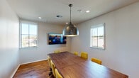 This single-story conference room combines contemporary design with warm, rustic elements to create a functional and inviting meeting space. The centerpiece is a substantial natural wood plank table surrounded by vibrant yellow upholstered chairs on casters, providing both comfort and a striking pop of color against the neutral white walls.

Warm hardwood flooring runs throughout, complementing the room's earthy aesthetic. A standout decorative feature is the large hammered-metal dome pendant light suspended by a chain from the ceiling, adding an industrial-chic character to the space. Recessed lighting supplements the pendant fixture, ensuring well-distributed illumination throughout.

Two wall-mounted flat-screen televisions are positioned at opposite ends of the table, making the room well-equipped for presentations and video conferencing. Multiple windows along the walls allow natural light to fill the space while offering views of the surrounding exterior.

A sleek, dark-framed frosted glass door provides privacy while maintaining a modern aesthetic and connecting the conference room to adjacent areas visible beyond — including a larger open space with exposed wood beam ceilings. A framed piece of colorful wall art adds a finishing decorative touch, reinforcing the room's blend of professional functionality and contemporary style.