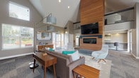 Located at 14320 SW Teal Blvd in Beaverton, Oregon, this single-story community clubhouse is designed with a contemporary mid-century modern aesthetic that balances style and functionality. The space is anchored by dramatically vaulted ceilings that create an airy, open atmosphere throughout the main lounge area. A defining architectural feature is the floor-to-ceiling wood-clad accent wall, which houses a sleek linear fireplace below and a large wall-mounted flat-screen television above, serving as a natural focal point for the room.

The main lounge is furnished with a generous sectional sofa, modern swivel accent chairs in neutral tones, and warm wood side tables, all arranged atop a layered area rug. Teal accent pillows echo the Pacific Northwest color palette and add a welcoming pop of color. Natural light filters in through multiple windows and glass doors, offering views of the surrounding outdoor patio and landscaped grounds.

Adjacent to the lounge, a dedicated workspace area features a long wall-mounted counter with bar-height seating, ideal for individual work or study. A separate, intimate seating nook with tufted red armchairs provides a quieter retreat. The fully equipped galley kitchen includes white shaker cabinetry, clean white countertops, stainless appliances, and direct access to an outdoor patio area, making the space well-suited for community gatherings and events.