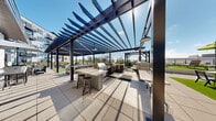 Located at 120 Summit St in Duluth, Minnesota, this expansive outdoor amenity space offers a thoughtfully designed, multi-zone courtyard and terrace environment spanning a generous footprint. The space is defined by its contemporary architectural aesthetic, featuring bold dark-metal pergolas with slatted louvered roofing that provide partial shade over fully equipped outdoor kitchen and grilling stations outfitted with stainless steel appliances and bar-height seating. Raised concrete planters filled with seasonal ornamental grasses and flowering perennials add vibrant natural texture throughout the space. A lush synthetic turf putting green and lawn area provides recreational space, complemented by clusters of colorful outdoor dining sets beneath bright umbrellas. The surrounding multi-story building, clad in a mix of corrugated metal panels and warm wood accents, frames the courtyard and adds a modern industrial character to the setting. Wide-format concrete pavers create clean, accessible pathways connecting each zone seamlessly. The open-air layout benefits from abundant natural light and sweeping views of the tree-lined Duluth skyline. Whether used for social gatherings, outdoor dining, or leisure, this well-appointed outdoor space delivers a premium amenity experience suited for year-round enjoyment in all its varied activity areas.