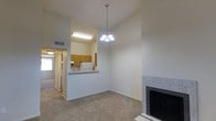 This single-floor, one-bedroom, one-bathroom unit offers a well-organized layout that maximizes its compact footprint. The main living area is carpeted and anchored by a tile-surround fireplace, adding a warm and inviting focal point to the room. A decorative chandelier hangs overhead, complementing the vaulted ceiling that lends a sense of added vertical space and openness.

The kitchen is positioned adjacent to the living area and features a pass-through counter that creates a connected, open feel between the two spaces. Warm wood-toned cabinetry lines the upper walls of the kitchen, providing ample storage, while a white refrigerator and overhead fluorescent lighting contribute to a clean, functional workspace.

A hallway visible from the main living space leads toward the bedroom and bathroom, maintaining a straightforward and efficient flow throughout the unit. Natural light enters through a window visible in the background, brightening the interior. The neutral color palette throughout — from the beige carpet to the white walls — creates a versatile backdrop suited to a variety of furnishing styles.