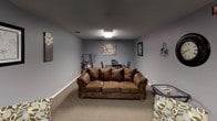 Located at 4700 S Baha Ave in Sioux Falls, South Dakota, this single-floor interior space offers a welcoming and functional layout designed for comfortable gathering and relaxation. The space is finished with soft gray walls and textured ceilings throughout, lending a calm, cohesive aesthetic from room to room.

The main lounge area is generously sized and anchored by a plush, brown upholstered sofa complemented by patterned accent chairs and glass-top side tables. A wall-mounted flat-screen television sits across from the seating arrangement, making the area well-suited for both casual conversation and entertainment. Flush ceiling light fixtures provide steady, even illumination across the carpeted floors.

Adjacent to the lounge is a dedicated dining area featuring a dark-toned table with matching chairs, ideal for group meals or meetings. Decorative artwork adorns the walls throughout, including floral canvas prints and framed photographs that add warmth and visual interest to the space.

A set of double doors connects to an adjoining hallway or room, suggesting additional functional space beyond the main lounge. The overall layout flows naturally between the seating and dining zones, making the space versatile and practical. Neutral carpet flooring ties all areas together, contributing to the space's understated, comfortable atmosphere.