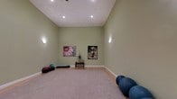 This tranquil meditation room is thoughtfully designed to foster a sense of calm and focus. The space is defined by soaring vaulted ceilings that create an open, airy feel, while the soft sage-green walls contribute to a soothing, grounded atmosphere. Recessed lighting is complemented by subtle wall-mounted accent lights that cast a gentle, warm glow throughout the room, making it ideal for both daytime and evening practice. The flooring transitions from warm hardwood along the perimeter to a large, plush area rug at the center, providing a comfortable surface for seated or floor-based practice. Along the back wall, a small console table serves as a subtle focal point, adorned with a decorative plant, while two art pieces — a vibrant floral canvas and a contemplative Buddha portrait — add visual depth and reinforce the room's mindful character. Exercise and stability balls are neatly arranged in one corner, and meditation cushions and mats along the opposite wall underscore the room's versatility for yoga, stretching, and breathwork. Clean white baseboards and a minimalist layout keep the space uncluttered, allowing for an environment fully conducive to reflection and inner stillness.