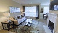 This single-floor residence offers a thoughtfully designed layout that balances comfort and style. The living area serves as the centerpiece of the home, featuring large windows dressed with elegant gray curtain panels and white blinds that flood the room with natural light. Neutral wall tones and crown molding lend a refined architectural touch throughout the space.

The living room is generously proportioned, accommodating a plush white sofa accented with patterned throw pillows and anchored by a bold geometric area rug in earthy tones. A sleek glass-and-metal coffee table sits at the center, adding a contemporary contrast to the warm, neutral palette. Decorative wall art and a tasteful floor lamp contribute to the polished ambiance.

Soft carpeting flows throughout the space, enhancing the warm and welcoming atmosphere. A glimpse into the adjacent area reveals a dining space, suggesting an open and connected floor plan that supports easy movement between living and dining zones. The overall aesthetic is clean, modern, and carefully curated, making this residence an inviting and functional living environment suitable for a variety of lifestyles.
