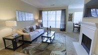 This single-floor residence offers a thoughtfully designed layout that balances comfort and style. The living area serves as the centerpiece of the home, featuring large windows dressed with elegant gray curtain panels and white blinds that flood the room with natural light. Neutral wall tones and crown molding lend a refined architectural touch throughout the space.

The living room is generously proportioned, accommodating a plush white sofa accented with patterned throw pillows and anchored by a bold geometric area rug in earthy tones. A sleek glass-and-metal coffee table sits at the center, adding a contemporary contrast to the warm, neutral palette. Decorative wall art and a tasteful floor lamp contribute to the polished ambiance.

Soft carpeting flows throughout the space, enhancing the warm and welcoming atmosphere. A glimpse into the adjacent area reveals a dining space, suggesting an open and connected floor plan that supports easy movement between living and dining zones. The overall aesthetic is clean, modern, and carefully curated, making this residence an inviting and functional living environment suitable for a variety of lifestyles.
