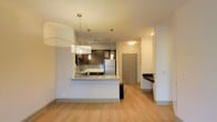 This single-floor space features an open-concept design that seamlessly connects the living area to the kitchen. The kitchen is equipped with dark espresso cabinetry, a stainless steel refrigerator, and a gas range, complemented by a tiled backsplash and granite countertops along the peninsula. The peninsula serves as both a workspace and casual dining area, with pendant lighting above adding a modern touch. A track lighting system in the kitchen provides ample task lighting, while a large contemporary pendant light anchors the living area. Light hardwood flooring runs throughout the space, contributing to a warm and cohesive aesthetic. Near the entryway, a built-in shelf or desk nook offers a practical workspace or storage solution. The unit entry door is visible from the main living area, reinforcing the efficient and straightforward layout. Neutral wall tones and clean lines throughout give the space a bright, contemporary feel that is both functional and visually appealing.
