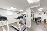 Located at 7700 Eastern Avenue Northwest in Washington, DC, this fitness center offers a fully equipped exercise room designed to support a well-rounded workout routine. The space features a clean, minimalist aesthetic with bright white walls and durable dark concrete flooring throughout, creating a functional and focused environment for physical activity.

The room is illuminated by recessed skylight-style ceiling panels that provide consistent, even lighting across the entire floor. A wall-mounted air conditioning unit ensures climate control year-round, while small windows along one wall allow for a modest amount of natural light. Structural columns divide the space into functional zones without impeding usability.

The equipment selection is comprehensive for a compact facility. Cardio offerings include multiple treadmills and elliptical trainers, as well as a stationary bike. For strength and conditioning, the room is stocked with a full rack of dumbbells in varying weights, a medicine ball rack, a stability ball, a mini trampoline, and a workout bench. Wall-mounted flat-screen televisions are positioned for easy viewing during exercise. Built-in cabinetry provides additional storage. Safety features such as a fire alarm, exit signage, and posted facility guidelines are also present, reflecting a well-maintained and regulation-compliant exercise environment.