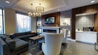 This beautifully appointed lounge space exudes contemporary elegance through a carefully curated blend of textures, materials, and design elements. The ceiling immediately draws the eye with its intricate geometric patterned relief detail, complemented by a pair of stunning multi-bulb chandelier fixtures that cast warm, ambient light throughout the room. Bold botanical-patterned wallcoverings in deep navy and grey tones anchor the walls, adding dramatic visual depth alongside rich walnut wood paneling. At the heart of the space, a sleek linear fireplace serves as the focal point, framed by the wood paneling and topped with a wall-mounted flat-screen television. Built-in display niches with white trim provide tasteful spots for decorative accents. The seating area is thoughtfully arranged with a plush dark sectional sofa, a rustic wood-and-metal coffee table, and elegant high-back accent chairs upholstered in soft neutral tones with gold nail-head trim detailing. A tufted ottoman in muted blue adds a refined pop of color. Large windows allow natural light to filter through sheer drapery, brightening the space and offering a sense of openness. The overall ambiance is polished, inviting, and meticulously designed.