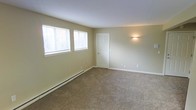 This single-floor residential space offers a clean, functional layout designed for comfortable everyday living. The interior is finished with warm, neutral-toned walls that create a calm and inviting atmosphere throughout. Wall-to-wall carpeting in a classic beige tone runs underfoot, adding warmth and softness to the living area. Natural light filters in through multiple windows fitted with horizontal blinds, allowing for flexible light control while maintaining a bright and airy feel. A classic six-panel white door provides access between rooms, complementing the crisp white baseboards and trim that frame the space neatly. A wall-mounted light fixture adds a subtle accent, offering additional illumination as needed. The baseboard heating system along the perimeter wall ensures efficient climate control during cooler months. The overall layout is open and uncluttered, lending itself well to a variety of furniture arrangements. The consistent finishes and understated design elements throughout the space create a cohesive and practical living environment suitable for a range of occupants.