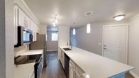 This well-appointed 2-bedroom, 2-bathroom single-floor residence offers a comfortable and modern living experience. The kitchen serves as a central highlight, featuring an expansive quartz countertop island with an undermount sink and a high-arc pull-down faucet, providing generous prep and dining space. White shaker-style cabinetry lines the walls, complemented by stainless steel appliances including a refrigerator, range, and dishwasher. Stylish pendant lighting hangs above the island, while track lighting illuminates the cooking area, creating a bright and functional workspace. Crown molding adds a refined architectural detail throughout. Dark hardwood-style flooring runs through the main living areas, offering a rich contrast to the light-toned walls and cabinetry. Natural light filters in through windows visible down the hallway, contributing to an open and airy atmosphere. The layout flows efficiently across a single floor, connecting the kitchen and living areas seamlessly. With two full bathrooms and two bedrooms, this residence is designed to accommodate comfortable everyday living with a clean, contemporary aesthetic.