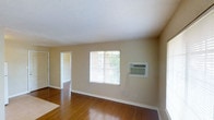 This single-floor residential space offers a clean and functional layout designed for comfortable everyday living. The main living area is characterized by rich hardwood flooring that extends throughout, adding warmth and a sense of continuity to the space. Neutral beige walls provide a versatile backdrop, making the area easy to personalize and decorate to any style.

A prominent window in the main room allows natural light to flow in, brightening the interior and offering a view of the surrounding greenery outside. A wall-mounted air conditioning unit is conveniently positioned beside the window, ensuring climate control throughout the warmer months. White baseboards provide a crisp contrast against both the walls and the flooring, contributing to a tidy, well-finished appearance.

The textured white ceiling adds a subtle architectural detail that complements the overall aesthetic. An open doorway leads into an adjacent room, also finished with matching hardwood floors, suggesting a cohesive design carried throughout the space. The layout feels open and connected, allowing for easy movement between areas. Overall, this space presents a straightforward, move-in-ready environment with practical features and a welcoming ambiance.