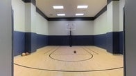 This indoor basketball court offers a well-designed, self-contained athletic space ideal for practice and recreational play. The court features a classic hardwood floor with clearly marked court lines, including a three-point arc and key, providing a professional playing surface that is both durable and high-performing. A regulation-height basketball hoop with a clear acrylic backboard is mounted at center court, offering an authentic game experience. Safety is a clear priority, as the lower half of all surrounding walls is lined with dark navy padded panels, protecting players during intense gameplay. The upper walls are painted in a clean off-white tone, accented by a bold dark horizontal band that adds a sharp, sporty aesthetic. Overhead, large recessed flat-panel LED light fixtures are evenly distributed across the ceiling, flooding the court with bright, consistent illumination to ensure excellent visibility throughout. The ceiling height is generous, providing ample clearance for jump shots and overhead play. The overall design balances functionality with a polished, professional appearance, making this a standout amenity suited for athletes of all skill levels seeking a premium indoor court experience year-round.