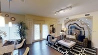 This single-floor space showcases a thoughtfully designed living area that balances contemporary style with comfortable functionality. The room is anchored by rich, dark hardwood flooring that runs throughout, creating a warm and cohesive foundation. Overhead, a sleek track lighting system provides adjustable, directed illumination across the space, complementing the natural light that pours in through a set of white-framed French doors dressed with sheer, patterned curtains and horizontal blinds.

A standout decorative feature is the large-scale agate geode wall art piece that commands attention on one side of the room, adding an artistic and dramatic focal point. The living area is furnished with a dark upholstered sofa accented with decorative pillows, a white rectangular coffee table, and a patterned accent chair, all arranged atop a bold geometric black-and-white area rug. A tall indoor plant in the corner adds a natural, organic element to the space. Warm yellow-toned walls contribute to an inviting ambiance, while the overall design reflects a modern aesthetic with distinctive, curated details throughout.