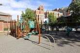 This outdoor playground is a well-appointed recreational space designed to encourage active play in a secure, carefully landscaped environment. The perimeter is defined by a sturdy metal fence interwoven with climbing ivy and anchored by classic brick columns, creating a welcoming yet enclosed boundary that ensures safety. Entry points are clearly marked, with gravel-lined pathways leading into the play area.

The playground surface is covered in rubberized gravel, providing impact-absorbing cushioning throughout the space. A variety of modern play structures occupy the area, including a multi-level climbing apparatus finished in teal and deep red tones, complete with staircases, railings, and elevated platforms. Freestanding curved climbing arches in a warm wood-toned finish add sculptural interest, while a hand-pump-style interactive play element and suspended disc swings offer additional sensory engagement.

Mature trees frame the space, casting dappled shade across the play area on sunny days and contributing to a pleasant, park-like atmosphere. Brick buildings are visible in the background, suggesting an urban or campus setting. The overall design balances functionality, safety, and aesthetic appeal, making it a thoughtfully designed space suitable for a range of ages and play styles.